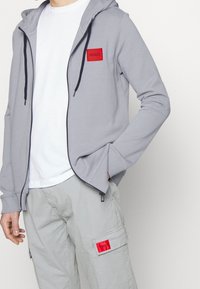 Sudadera gris con cremallera y parche de logo rojo "HUGO", capucha con cordones y cremallera azul marino. Combinada con pantalones cargo de color gris claro que presentan un parche de logo rojo.