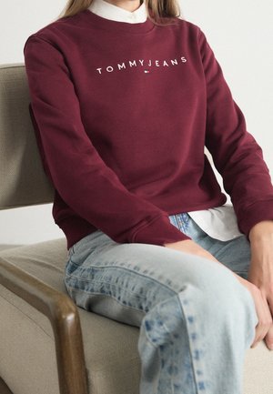 Persoon zittend, gekleed in een bordeauxrode Tommy Jeans trui over een wit shirt en lichtblauwe spijkerbroek, op een beige fauteuil.