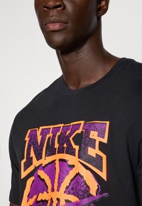 Sort bomulds T-shirt med et stort "NIKE" logo i orange og lilla samt et basketball grafik. Rund halsudskæring og korte ærmer.