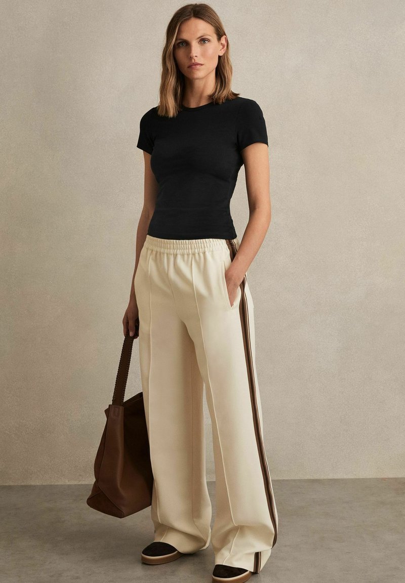 Modellen står mot en strukturerad beige bakgrund, klädd i en figursydd svart t-shirt och krämfärgade vida byxor, med en brun handväska i handen.