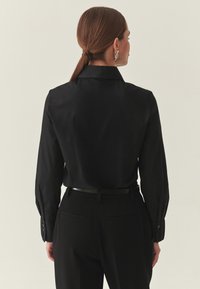 Fekete hosszú ujjú gombos ing hegyes gallérral és sima textúrával, fekete nadrággal és vékony övvel párosítva.