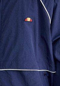 Marinejakke med et glat, letvægtigt materiale. Har hvid piping og et lille broderet logo i rød, gul og blå på brystet.