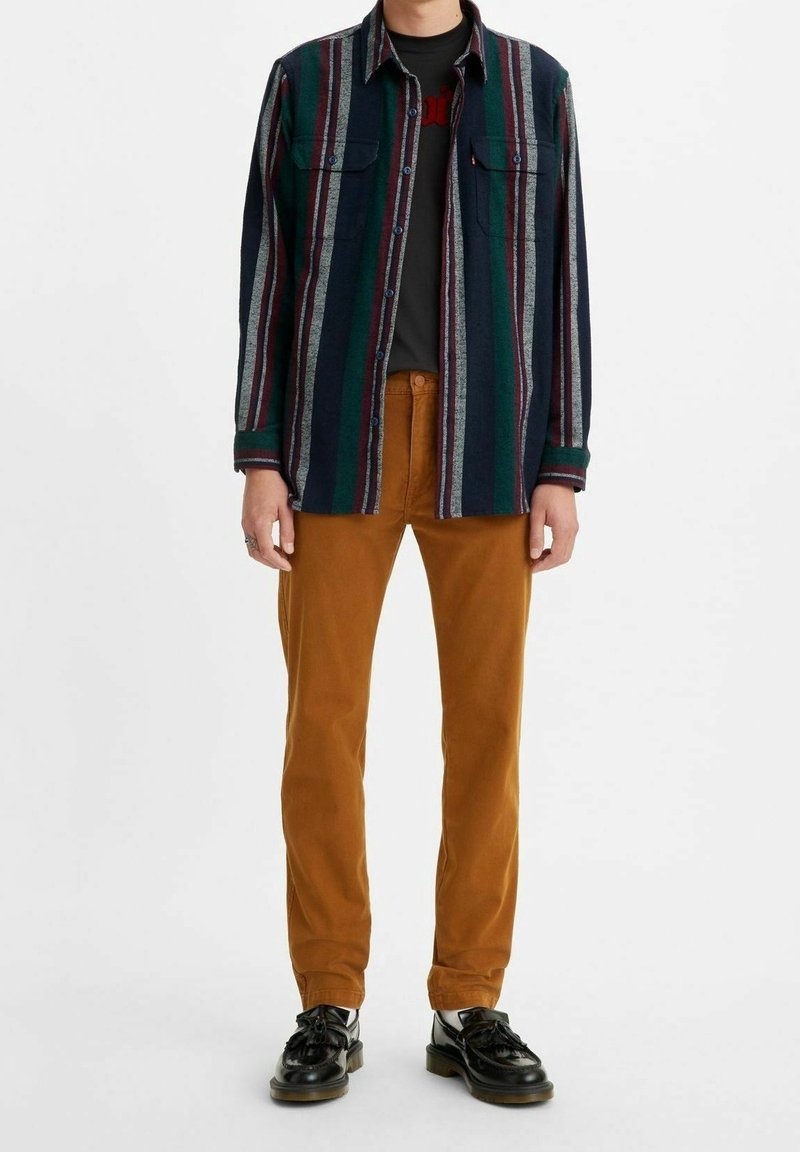 Personne portant une chemise boutonnée à rayures foncées, un t-shirt graphique noir, un pantalon couleur moutarde et des chaussures en cuir noires, debout sur fond blanc.