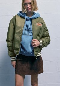 Olivevihreä pommittaja, jossa on valkoiset ja laivaston siniset raidat hihansuissa, ja siinä on oranssi brodeerattu "Tigers" -patch, kerroksittain vaaleansinisen hupparin ja ruskean mokkahihaisen hameen päällä.