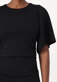 Black top with a round neckline and short, flared sleeves. Fabric is smooth with gentle texture and gathers at the waist.