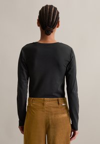 Persoon met een gevlochten haarbunt, gezien van achteren, die een zwart longsleeve shirt en een bruine corduroy broek draagt, tegen een effen achtergrond.
