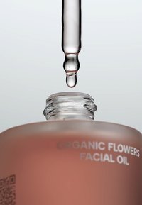 Szklana butelka olejku do twarzy z frosted różowym wykończeniem. Przezroczysta pipeta uwalniająca pojedynczą kroplę, oznaczona etykietą "Olej do twarzy z organicznych kwiatów".