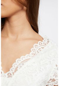 Haut en dentelle blanche avec un col scallopé. Motif floral complexe, texture douce et détails délicats le long des bords.
