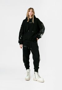 Conjunto de sudadera negra con capucha y joggers con acentos brillantes y similares a plumas en las mangas y piernas. Unos botines blancos gruesos con cordones completan el look.