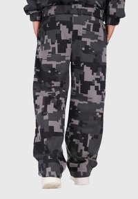 Osoba w luźnych czarno-szarych spodniach w cyfrowy kamuflaż z tylną kieszenią, z rękami w kieszeniach i białych sneakersach, stojąca na neutralnym tle.