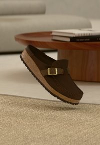 Καφέ μοκασίνι από σουέντ με φελλό σόλα και χρυσή αγκράφα, αιωρούμενο πάνω από μπεζ υφασμάτινο χαλί, κοντά σε ξύλινο τραπεζάκι καφέ με βιβλία.