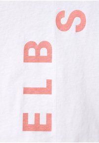 Valkoinen puuvillakangas, jossa on suuria, vaalean korallinvärisiä kirjaimia, jotka kirjoittavat "ELBS". Suunnittelussa on näkyvää ompelua sauman varrella.