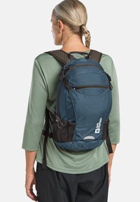 Teal rugzak met een textuur op de achterkant, twee zippered compartimenten, gaas zijzakken en een reflecterende accent, gedragen over een groen shirt.