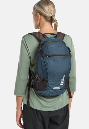 Türkise Rucksack mit strukturiertem Rücken, zwei Reißverschlussfächern, seitlichen Netz-Halterungen und einem reflektierenden Akzent, getragen über einem grünen Shirt.