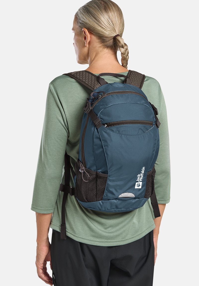 Teal rugzak met een textuur op de achterkant, twee zippered compartimenten, gaas zijzakken en een reflecterende accent, gedragen over een groen shirt.