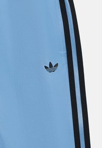 Pantalons de sport bleu clair avec texture verticale, bandes latérales noires et un petit logo Adidas noir. Fabriqué à partir d'un matériau synthétique lisse.