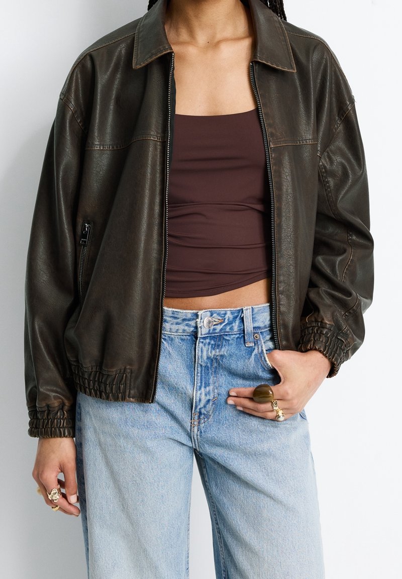Personne portant une veste en cuir marron foncé sur un crop top marron foncé, un jean taille haute bleu clair et plusieurs bagues aux deux mains.