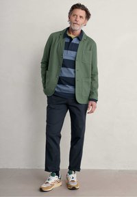 Chaqueta ligera verde sobre un polo a rayas en azul marino y gris, combinada con pantalones oscuros y zapatillas multicolores. Diseño sencillo, ajuste casual.