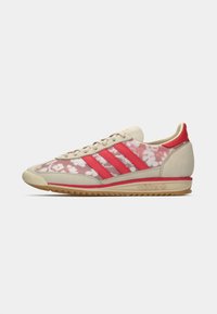 Adidas-sneakers med en lys beige semsket øvre, røde detaljer, blomstermønster og en gummisåle med stripete røde detaljer.
