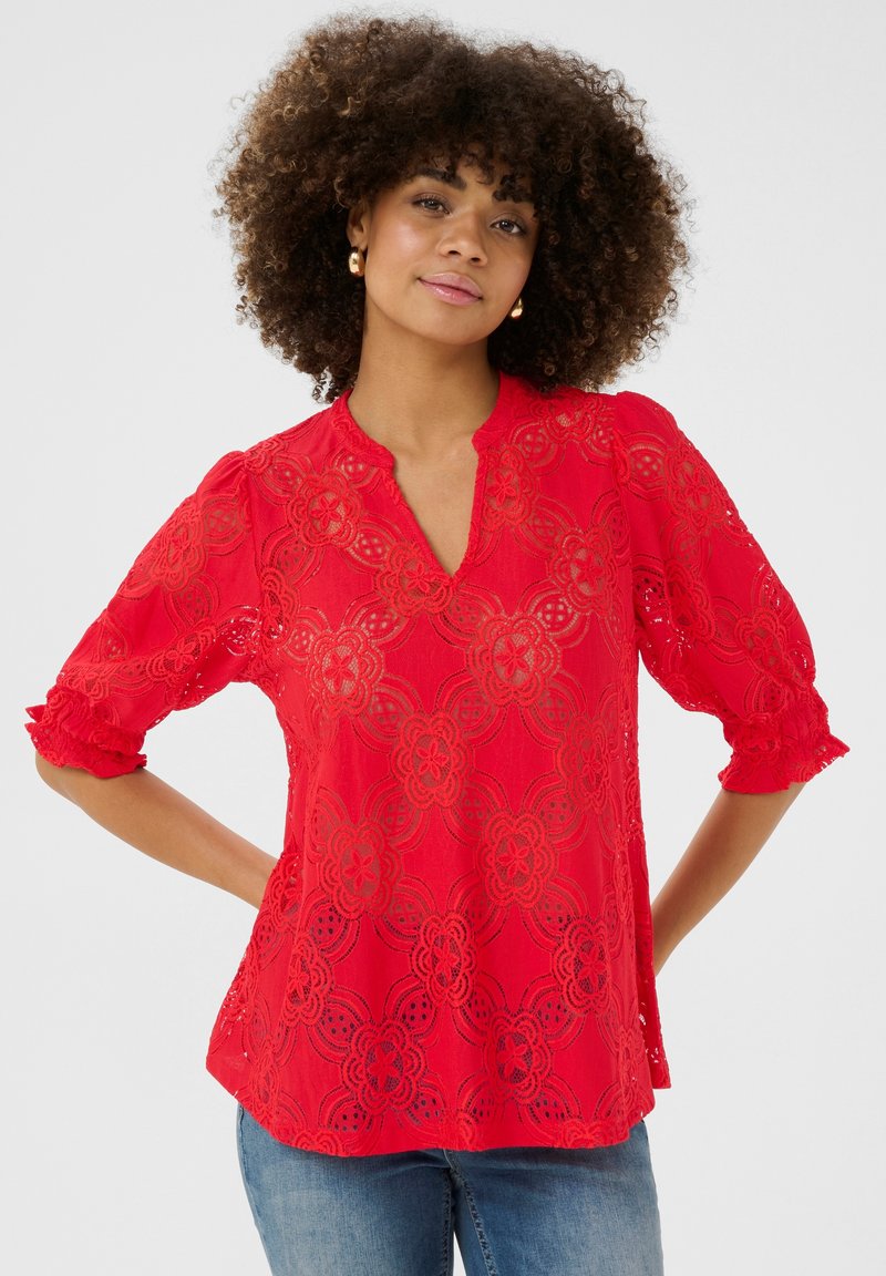 Top en dentelle rouge avec un col en V, orné de motifs floraux et de manches bouffantes aux bords volants. Associé à un jean bleu clair.