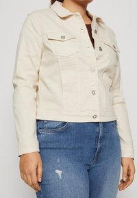 Jeansjacke in Cremefarbe mit geknöpftem Vorderteil, spitzem Kragen und zwei Brusttaschen. Verfügt über lange Ärmel und einen kurzen Schnitt.