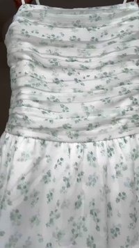 Biele šaty s jemným svetlozeleným kvetinovým vzorom, zdobeným vrškom s nariasením a tenkými ramienkami, visiace na tmavom pozadí.