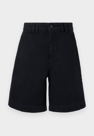 Nainen, joka on pukeutunut mustaan hihattomaan nappilistavestiin, vastaaviin polven mittaisiin shortseihin, ja pitää käsissään ruskeaa kudottua laukkua sekä vaaleita kudottuja kenkiä.