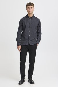 Chemise boutonnée gris foncé avec col, manches longues et boutons clairs contrastants, associée à un pantalon slim noir et des chaussures noires.
