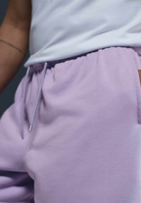 Pantalones de chándal de color púrpura claro con cintura elástica y detalles de cordón. La tela parece suave con una textura lisa. Se ve un bolsillo lateral.