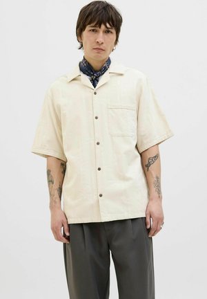 Short-sleeved, cream-colored button-up shirt with a textured pattern, single chest pocket, and brown buttons, paired with loose-fitting gray trousers.