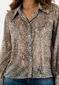 Camicia button-up leggera e trasparente in beige con motivo leopardato nero; presenta un colletto punta e una chiusura con un singolo bottone anteriore.