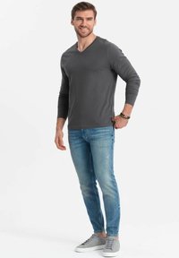 Graues langärmliges V-Hals-Shirt kombiniert mit hellblauen Jeans und grauen Sneakers. Das Model steht mit einer entspannten Haltung und einem leichten Lächeln.