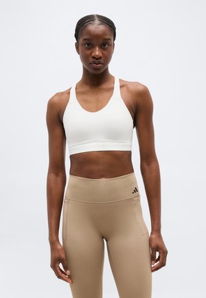 Γυναίκα με πλεγμένα μαλλιά που φοράει λευκό αθλητικό σουτιέν και αθλητικό κολάν μπεζ με ψηλή μέση, στέκεται μπροστά σε ένα απλό φόντο.