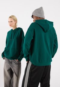 To personer har matchende mørk teal hettegensere med kengurulommer, kombinert med lysegrå og svarte bukser med hvite striper.