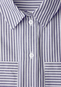 Gros plan sur le devant d'une chemise à rayures verticales bleues et blanches avec col, boutons blancs et deux poches plaquées à rayures horizontales.