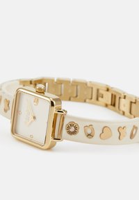 Quadratische Uhr mit goldfarbener Hardware, cremefarbenem Silikonarmband mit spielerischen goldenen Symbolen und einem goldumrandeten Zifferblatt im minimalistischen Design.