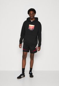 Černá mikina s kapucí se čtvercovým grafickým motivem na přední straně, v kombinaci s černými kraťasy s červenými akcenty a logem adidas. Černé tenisky dokončují celý vzhled.