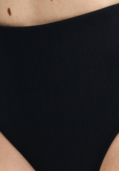 Κοντινό πλάνο ενός ατόμου που φορά μαύρο, ριμπ ύφασμα εσώρουχο ψηλής μέσης, με εμφανές σημάδι (ελιά) κοντά στη μέση.