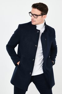 Cappotto doppiopetto blu navy in tessuto morbido con colletto alto, chiusura con bottoni e tasca sul petto con zip. Indossato sopra un dolcevita bianco.