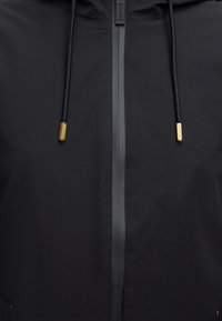 Zwarte jas met centrale waterdichte rits en zwarte trekkoorden met gouden metalen uiteinden gegraveerd met merknamen.