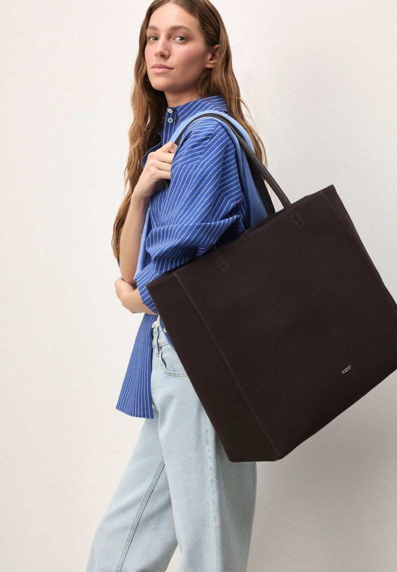 Sac fourre-tout noir en tissu robuste avec double poignée, forme carrée et détails de couture subtils. Le modèle porte une chemise à rayures bleues et un jean.