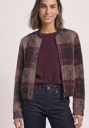 Femme souriante portant un pull violet sous une veste à carreaux zippée et un jean bleu foncé sur un fond clair.