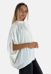 Haut blanc ample avec un col montant, des manches courtes et des détails découpés sur les côtés. Texture douce et matériau léger.