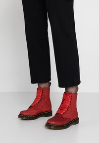 Röda glittriga ankelboots med bandspänne, runda tår och kraftig sula. Svarta byxor och nätstrumpor synliga, vilket framhäver skorna.