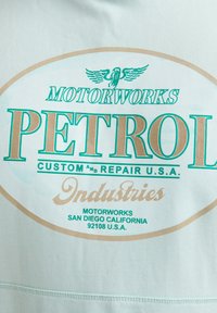 T-shirt bleu sarcelle clair avec un grand graphique ovale affichant "MOTORWORKS PETROL" en lettres vertes et dorées, avec un texte supplémentaire en dessous.