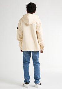 Sudadera beige con capucha y corte relajado, material de mezcla de algodón y puños de canalé, combinada con vaqueros de mezclilla azul claros y zapatillas negras y blancas.