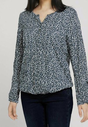 Vrouw die een blouse met lange mouwen draagt, zwart-wit abstract patroon, met knopen bij de hals en een donkere blauwe spijkerbroek.