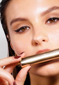 Guldig metallisk läppstift tub med texturerad yta. Närbild av händer som håller produkten, med mörkt bourgognefärgade naglar och slät hud.