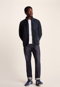 Dark green fleece jacket with a zip closure, standing collar, and chest pocket. Paired with dark denim jeans and blue sneakers.