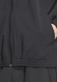 Μαύρο μπουφάν με φερμουάρ, ελαστική μέση και πλαϊνές τσέπες, φορεμένο με το ένα χέρι στην τσέπη, σε ένα άτομο.